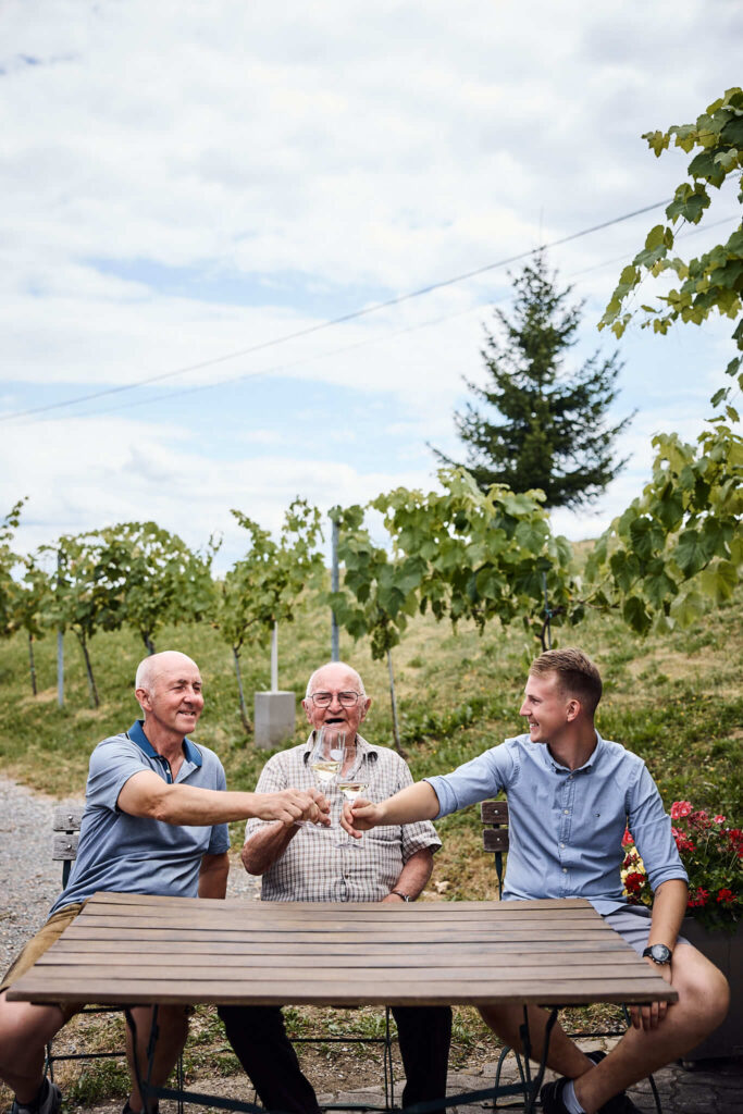 Matthias Strauss, Herbert Strauss stoßen mit Weingläsern an - Tisch im Weingarten